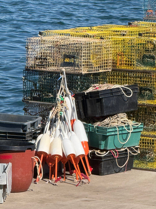 A photo of lobster traps and buoys stacked up on a dock with choppy waves in the background. 